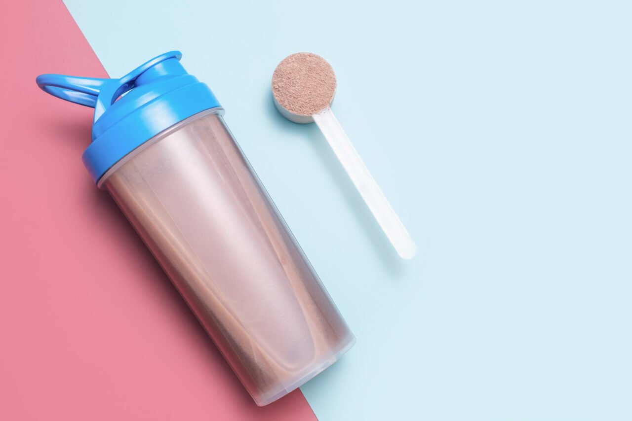 Bottle with cocktail and scoop with protein powder on blue and burgundy background close-up top view.